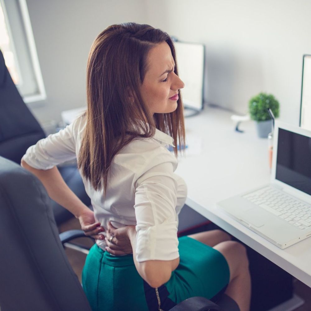 Woman experiencing back pain while sitting at a desk with a computer setup.
